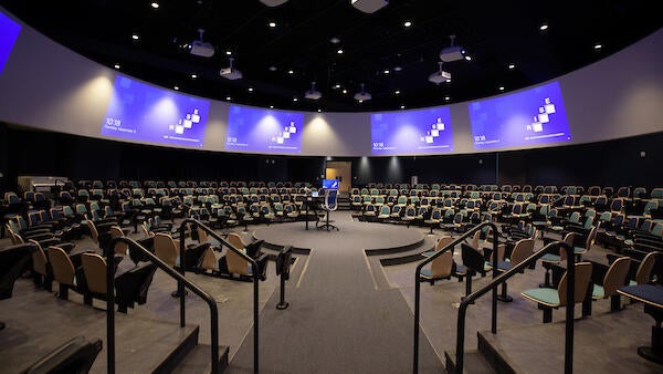A large lecture hall with seats set in a circular shape around the lecture podium with screens above surrounding the room.