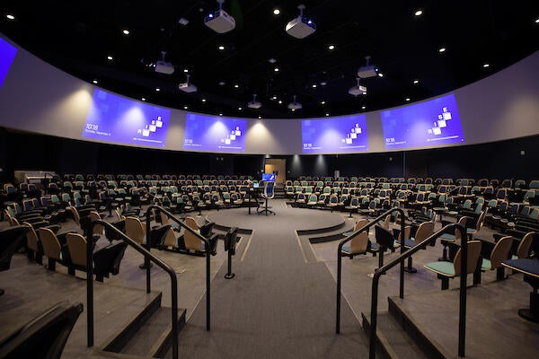 A large lecture hall with seats set in a circular shape around the lecture podium with screens above surrounding the room.